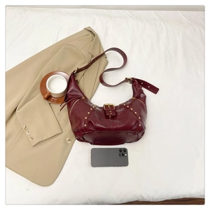 Détail d'un sac demi-lune rouge foncé avec rivets dorés sur fond de tissu blanc épuré