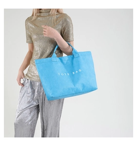 Detail of blue textured tote bag with TOTE BAG print worn by model in metallic outfit