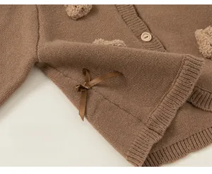 Detail of a brown knit cardigan featuring a plush bear patch and satin bow decoration