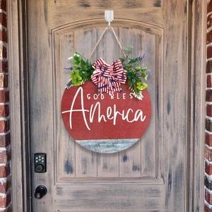 Round God Bless America wooden sign with flag bow and greenery on rustic door