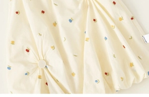Close-up of floral embroidery and pleated fabric detail on a cream cotton baby dress