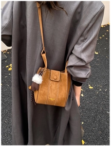 Brown suede bucket bag with contrast stitching and pom-pom charm shown on shoulder