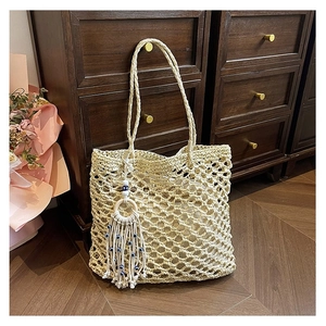 Beige hollow straw woven tote bag with Evil Eye tassel charm on a vintage wooden cabinet