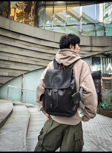 Street view of black faux leather backpack with flap cover and side pockets on a male model