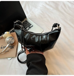 Detail of black patent leather crescent bag shown on a white bed with modern lifestyle props