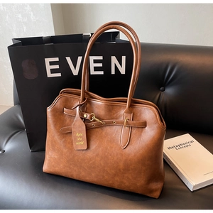 Brown leather tote bag featuring metal buckle strap and side tag on black leather sofa