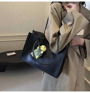 Detail of black pleated faux leather tote bag with green turtle plush charm on shoulder
