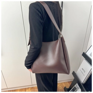 Deep brown leather tote bag with wide strap shown on shoulder in a minimalist room