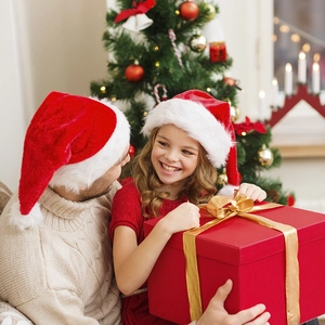 Father and daughter wearing plush red Christmas hats with festive gift box and tree background
