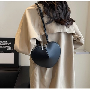 Dettaglio di una borsa a tracolla nera a forma di cuore indossata su un trench beige