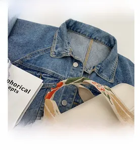Detail der cremefarbenen Canvas-Tragetasche mit bestickten Griffen auf einer Jeansjacke