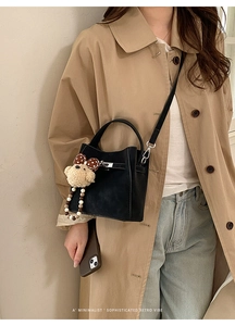 Detail of a black leather bucket bag styled with a bear charm on a beige trench coat