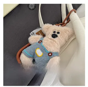 Close-up of a plush beige bear charm wearing blue knit overalls on a white bag