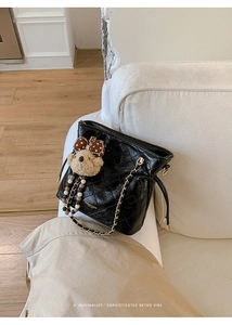 Close-up of black quilted bucket bag with gold chain strap and teddy bear charm on sofa