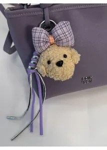 Close-up of a beige curly plush teddy bear charm with a purple plaid bow on a handbag