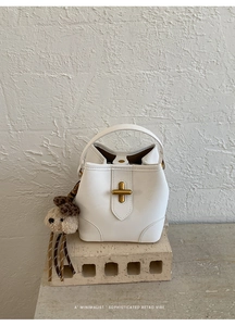 Détail du sac seau blanc avec boucle en croix dorée et breloque ours en peluche sur brique