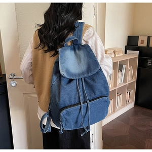 Detail of blue denim backpack with flap closure and drawstring worn by model indoors