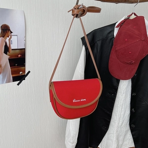 Red fabric saddle bag with brown leather trim and white typography hanging in a closet