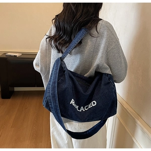 Model showcasing a large dark blue denim tote bag with white REPLACED lettering print
