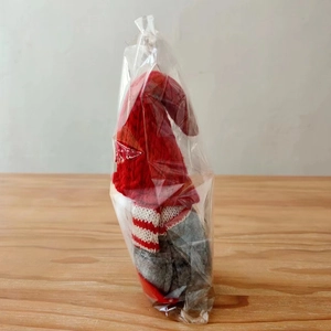 Detail of red and grey knitted Christmas gnome doll in transparent packaging on wood