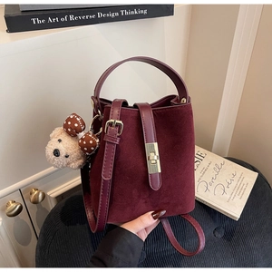 Détail d'un sac seau bordeaux mat avec boucle dorée et peluche sur un fauteuil sombre