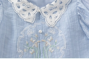 Detail of light blue pleated shirt showing lace collar, pearl button and floral embroidery
