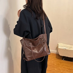 Large dark brown underarm tote bag in glossy leather worn by a model in a minimalist room