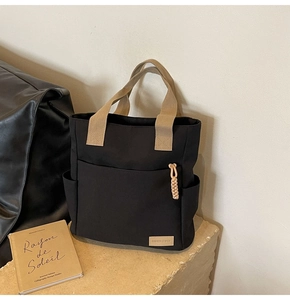 Detail of a large black canvas tote bag with khaki handles and front pocket on a stone base