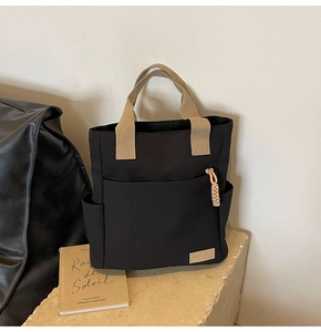 Close-up of black canvas tote bag with khaki handles and front pocket detail on stone base