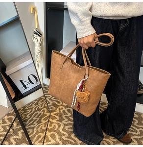 Deep brown tote bag with plush bear charm held by a model in a cozy indoor setting