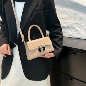 Elegant beige trapezoid handbag with gold T-clasp held by model in black blazer