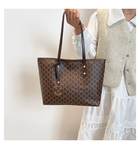 Detail of a brown tote bag with M-pattern print and a gold-tone metallic charm