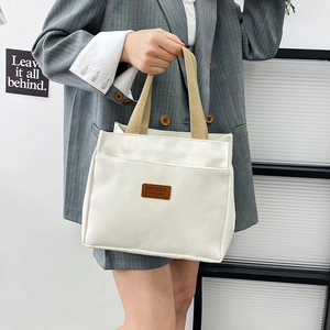 Minimalist beige canvas tote bag with brown sport label held by person in grey blazer