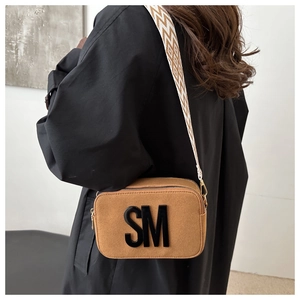 Brown camera bag with SM logo and wide geometric shoulder strap shown on black coat