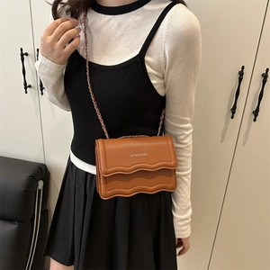 Brown leather crossbody bag with wavy flap design and chain strap shown on model