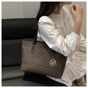 Detail view of large brown tote bag with M-pattern and gold charm on a leather sofa