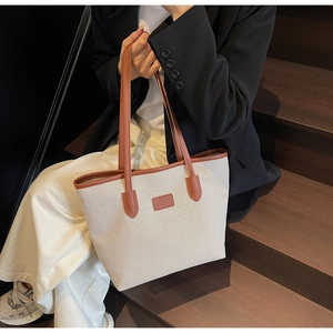 Detail of a minimalist cream tote bag with brown leather trim held by a seated model