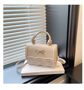 Detail of a beige handbag featuring a metal bow, scalloped edges, and knotted handle texture