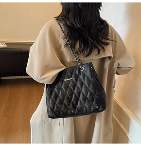 Detail of black quilted tote bag with silver chain strap worn over a beige trench coat