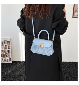 Détail du sac bandoulière bleu clair avec motif tressé et logo M doré sur manteau noir