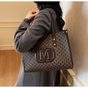 Close-up of brown tote bag featuring letter M monogram print and oval leather logo detail