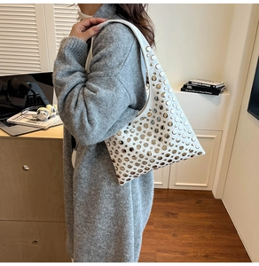 Detail of white half-moon bag with silver rivets worn over a grey knit cardigan