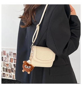 Detail of a creamy white flap crossbody bag with litchi texture and a brown plush monster charm