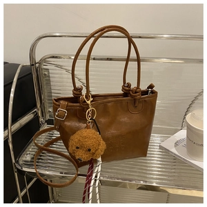 Deep brown tote bag with letter embossing and a cute teddy bear plush charm on a display shelf
