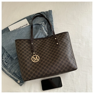 Detail of dark brown monogram tote bag with gold M-logo pendant on textured background
