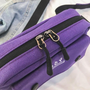 Detail view of the purple canvas messenger bag with dual metal zippers and logo detail