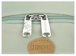 Detail view of the durable dual metal zippers and brown leather logo patch on the green backpack