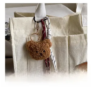 Close-up of a brown plush teddy bear keychain hanging on a white corduroy tote bag