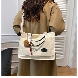 Detail of cream corduroy tote bag with fashion label and plush bear charm on broad strap
