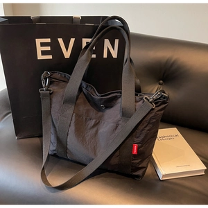 Close-up of black nylon tote bag on leather sofa with removable strap and red logo tag